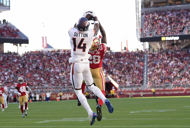 A player in white jersey number 14 leaps to catch a football while a player in a red jersey number 28 defends during a crowded stadium game.